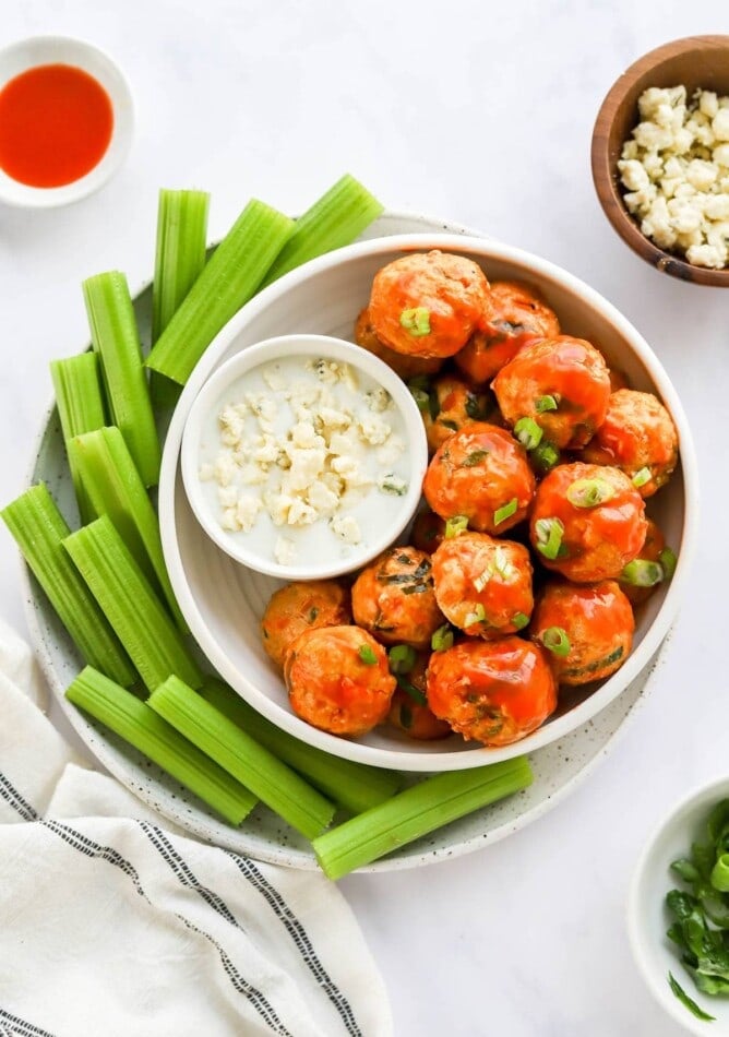 An overhead view of a bowl of buffalo chicken meatballs. A small bowl of blue cheese dip rests in the bowl with the meatballs and that bowl is on a plate surrounded by celery sticks.