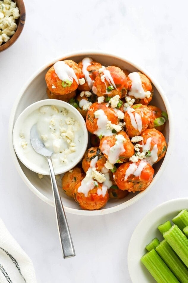 A bowl of buffalo chicken meatballs that have been drizzled with blue cheese. A bowl of blue cheese with a spoon resting in it is sitting inside the bowl of meatballs.