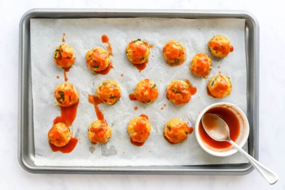 Fifteen uniform meatballs drizzled with buffalo sauce on a parchment lined sheet pan.
