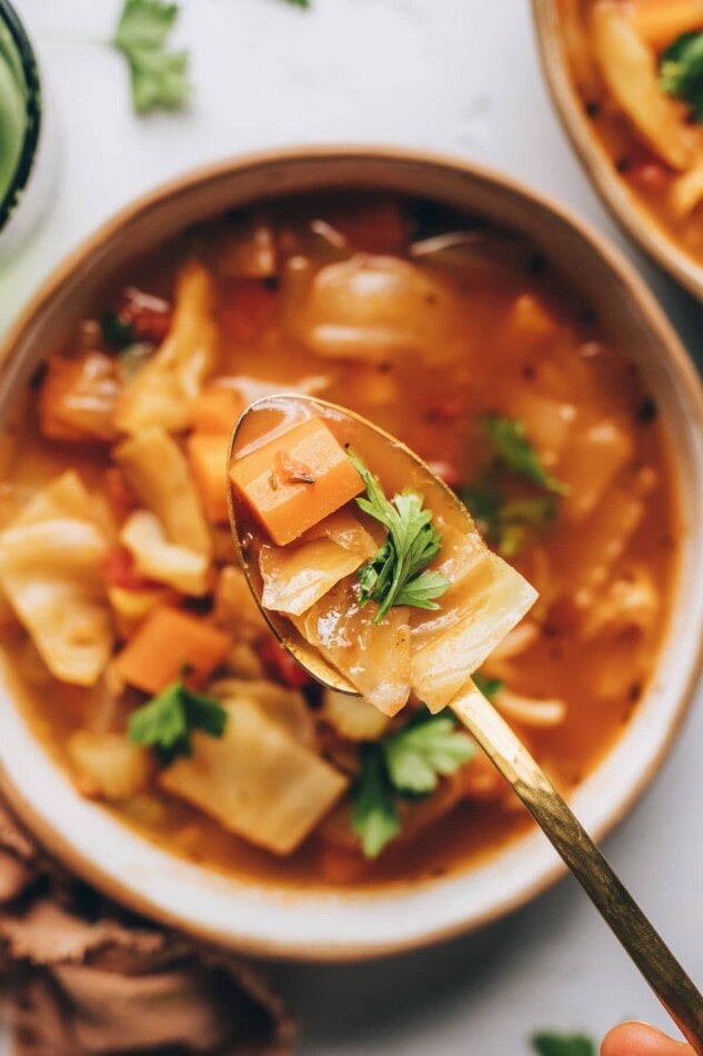 A spoon is holding up a bite of cabbage soup. The spoon is directly overtop of a bowl of the soup.