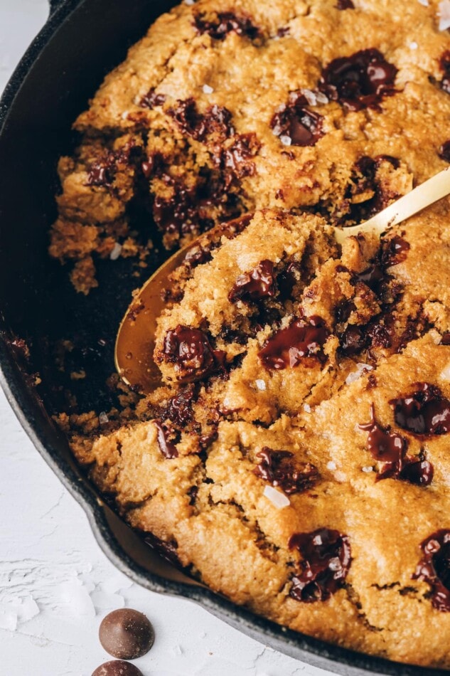 An overhead closeup photo of a chocolate chip cookie baked in a skillet. A golden spoon is scooping out a bite.