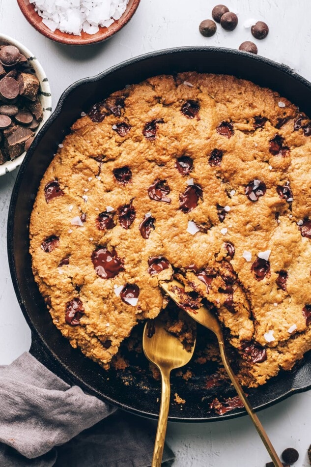 A cast iron skillet containing a chocolate chip cookie. Two spoons are removing a portion.