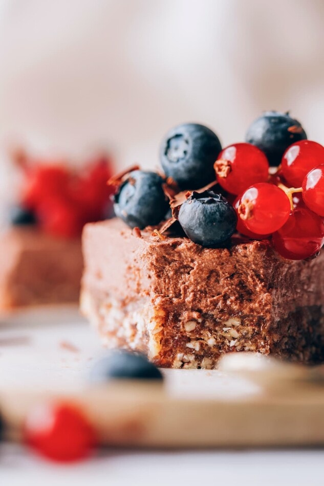 A close up of a slice of vegan chocolate tart. You can see the two layers of crust and filling. The slice is topped with blueberries and cherries.