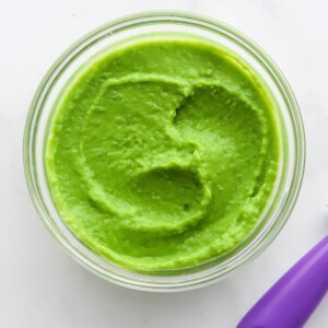 A small jar of pea puree. A purple spoon is cut out of the frame.