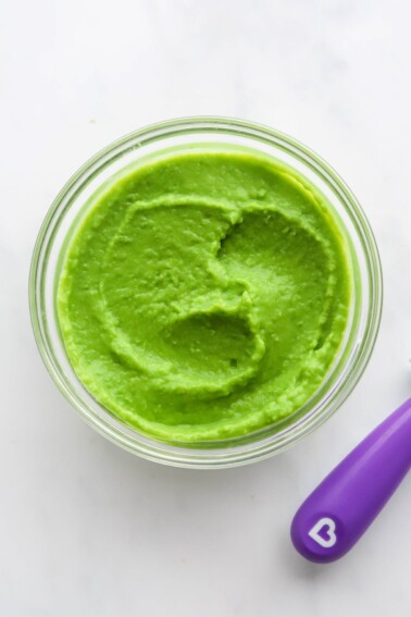 A small jar of pea puree. A purple spoon is cut out of the frame.