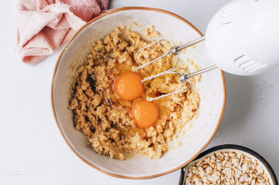 A medium mixing bowl with sugar, brown sugar and butter combined and fluffy. 2 Eggs and vanilla are being mixed in with a hand mixer.