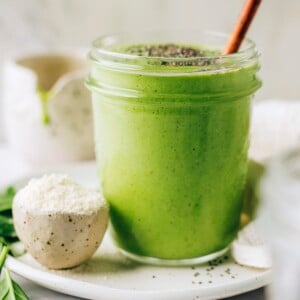 A mason jar containing green protein smoothie. The jar is resting on a plate with a small bowl of protein powder. The smoothie has been topped with chia seeds and a wooden straw sticks out of the top of the glass.