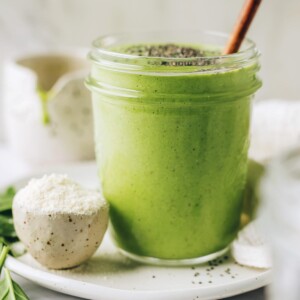 A mason jar containing green protein smoothie. The jar is resting on a plate with a small bowl of protein powder. The smoothie has been topped with chia seeds and a wooden straw sticks out of the top of the glass.