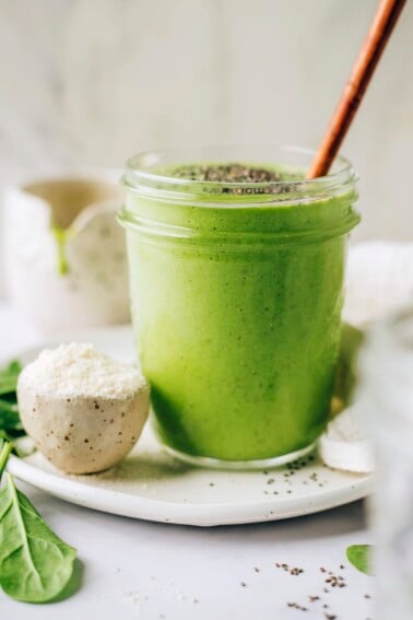 A mason jar containing green protein smoothie. The jar is resting on a plate with a small bowl of protein powder. The smoothie has been topped with chia seeds and a wooden straw sticks out of the top of the glass.