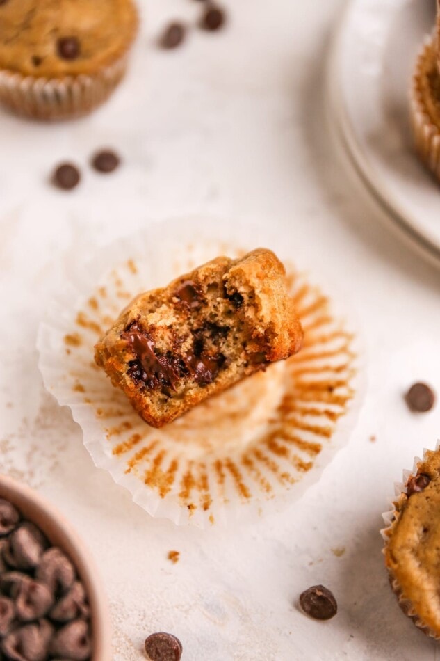 A muffin unwrapped from its paper lining. It is laying on its side on top of the lining with a bite removed.