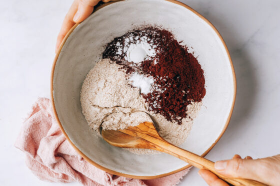 A wooden spoon is mixing together flour, cocoa powder, baking soda, baking powder and salt in a bowl.