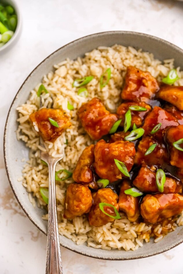 An overhead photo looking at a bowl of healthy orange chicken served over rice. A fork has a piece of chicken on it.