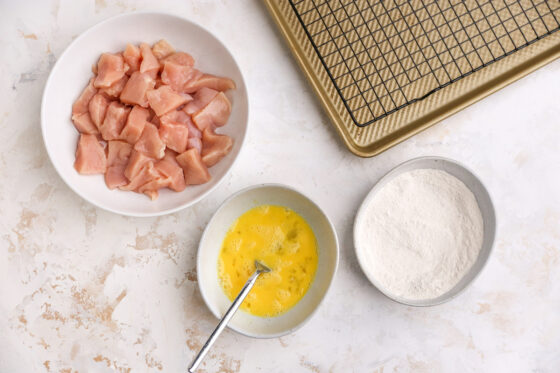 Chicken breast chunks in a bowl next to a bowl of egg wash and arrowroot powder and flour mixture.