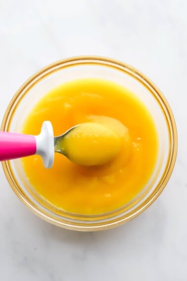 A small glass bowl containing mango puree. A small baby-sized spoon is lifting out a bite.