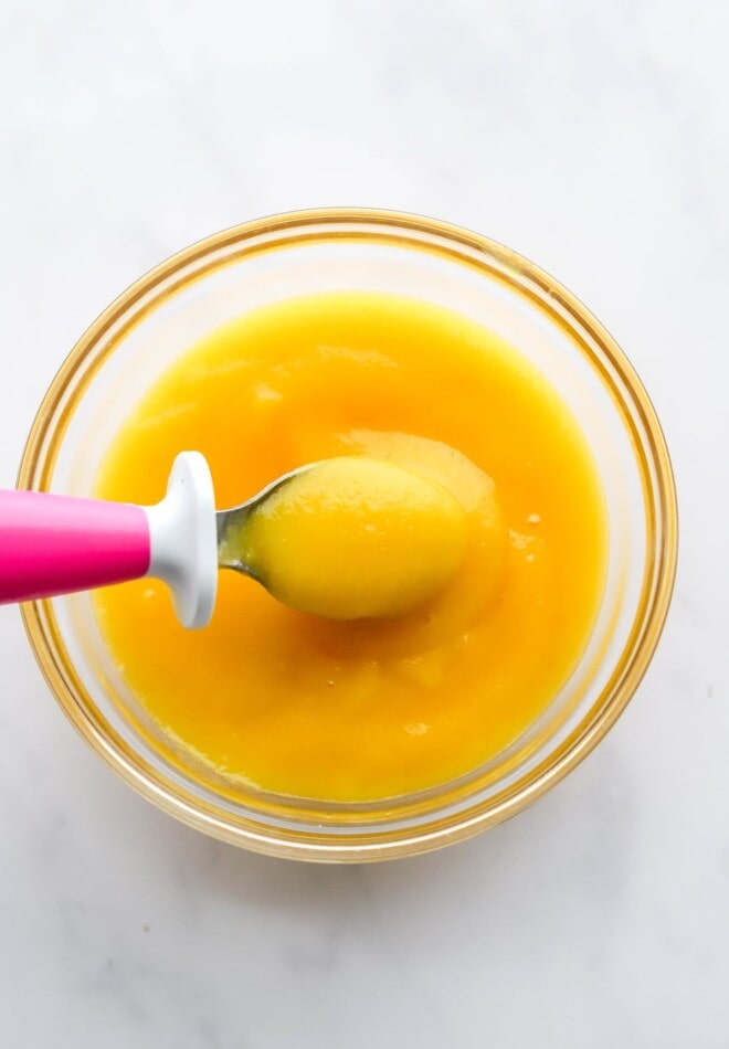 A small glass bowl containing mango puree. A small baby-sized spoon is lifting out a bite.