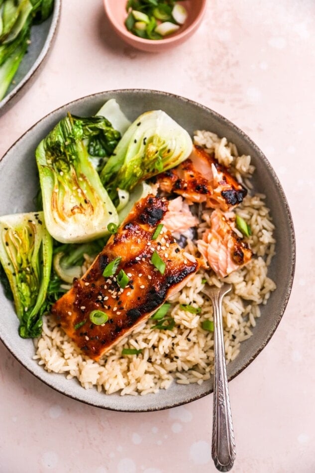 An overhead photo looking at a shallow bowl with a filet of miso salmon over a bed of brown rice with bok choy. A fork has separated some of the filet.