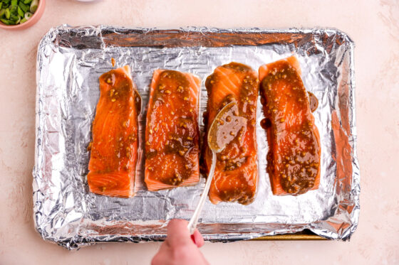 Four salmon filets on a baking sheet lined with foil. A spoon is being used to coat the salmon in the miso sauce mixture.