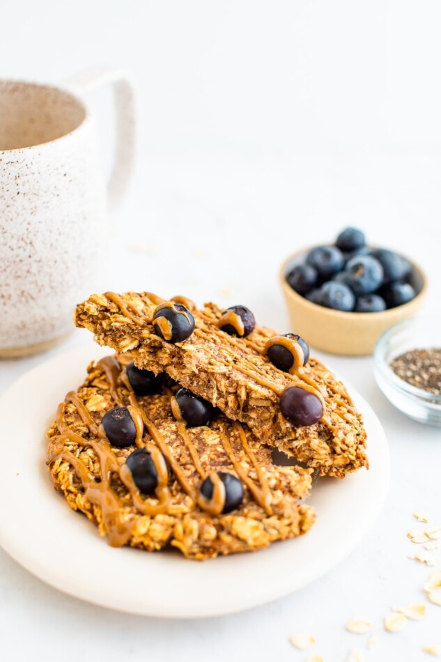 An oatmeal breakfast cookie on a plate that has been cut in half, the halves are resting on each other exposing the inside of the cookie. Behind the plate is a small container of chia seeds and blueberries as well as a mug of tea.