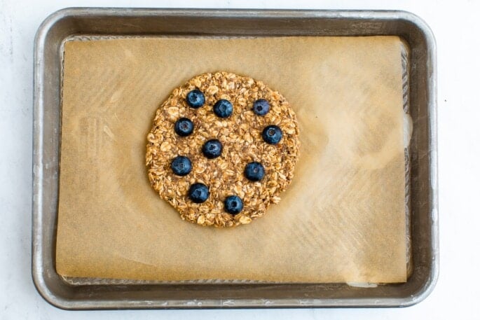 The oatmeal mixture has been poured onto a baking sheet lined with parchment paper and shaped into a circle. The top of the oatmeal mixture has been topped with 10 randomly placed blueberries.