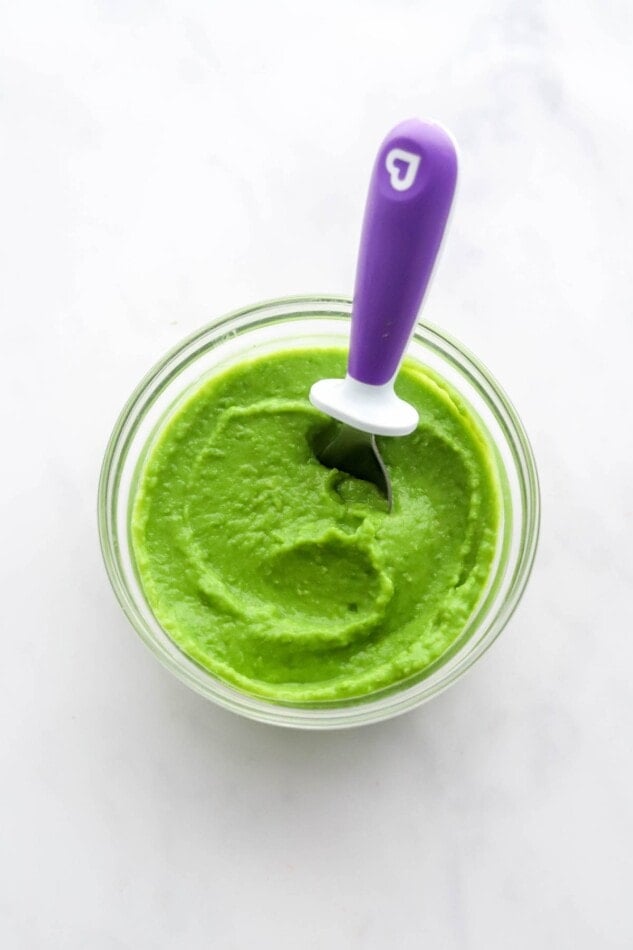 A small jar of pea puree. A purple baby spoon is resting in the jar.