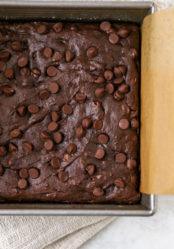 A baking pan lined with brown parchment paper containing protein brownies topped with chocolate chips.