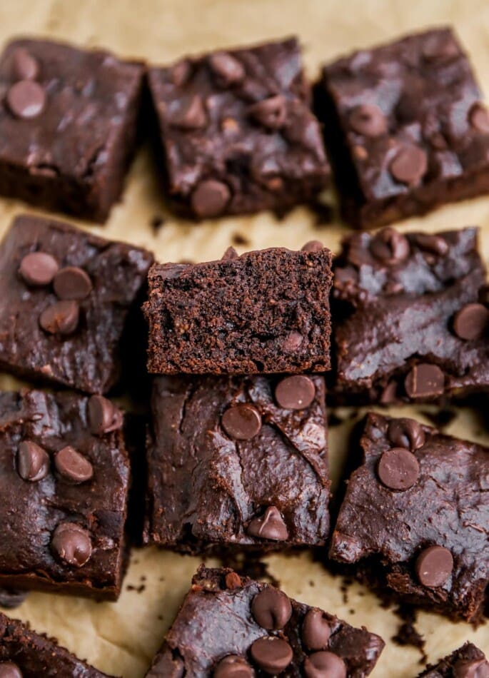 Squares of brownies on a sheet of brown parchment paper. The center brownie is resting against another brownie, exposing the inside.