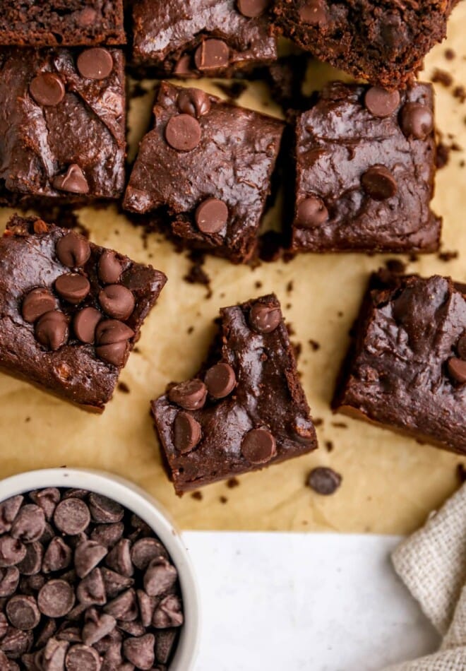 An overhead photo looking out a sheet of brown parchment paper with protein brownie squares on top. The center brownie has a bite taken out of it. There is a bowl of chocolate chips in the bottom left corner.