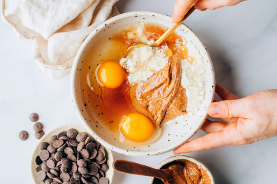 A mixing bowl containing almond butter, eggs, vanilla protein powder and maple syrup being mixed together.