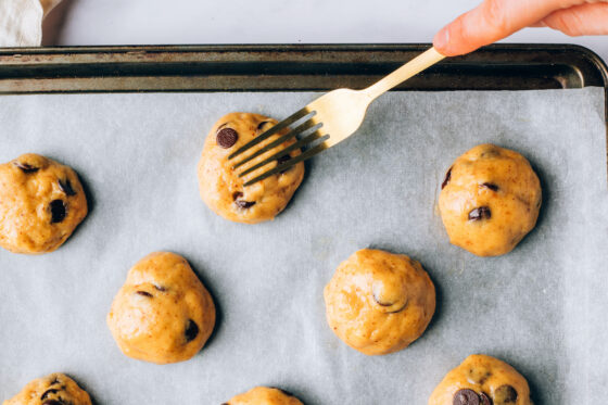 Portioned cookie dough balls on a parchment lined baking sheet. A fork is being used to flatten each cookie dough ball.