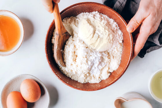 A wooden spoon mixing together flour, protein, baking powder and salt in a bowl.