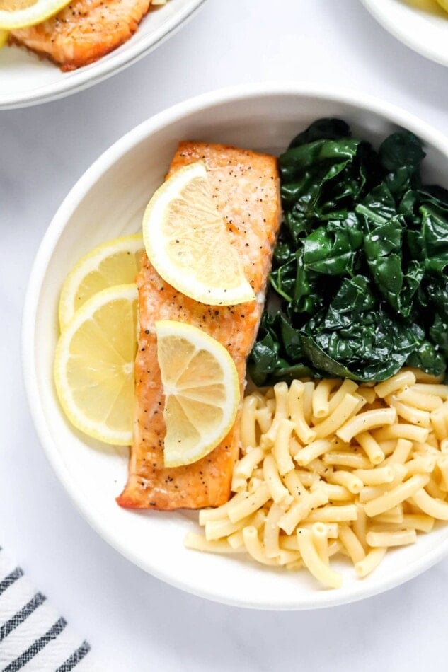 A bowl with macaroni and cheese, kale and a filet of salmon. Lemon slices top the salmon filet.