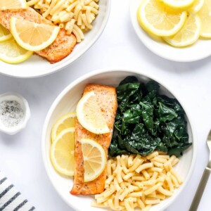 An overhead photo of a bowl with macaroni and cheese, kale and a filet of salmon. Lemon slices top the salmon filet.