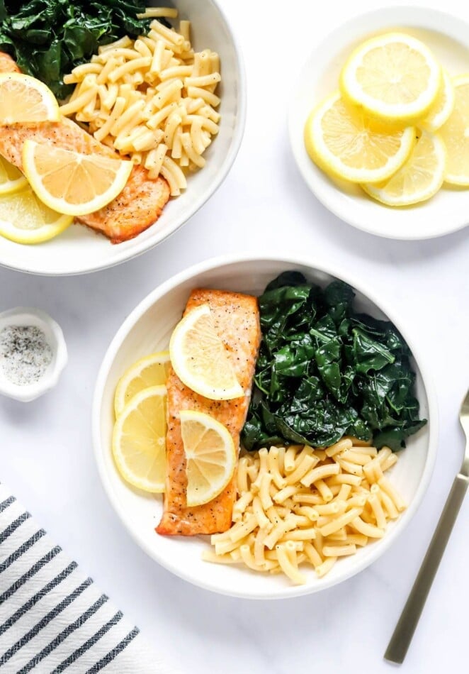 An overhead photo of a bowl with macaroni and cheese, kale and a filet of salmon. Lemon slices top the salmon filet.
