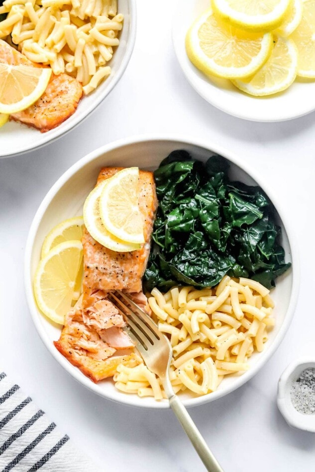 An overhead photo of a bowl with macaroni and cheese, kale and a filet of salmon. Lemon slices top the salmon filet and a fork is breaking up the salmon filet.