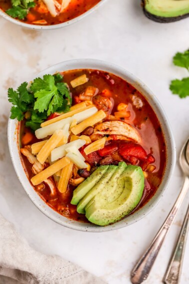 A bowl of chicken enchilada soup, topped with shredded cheese, avocado slices and fresh cilantro.