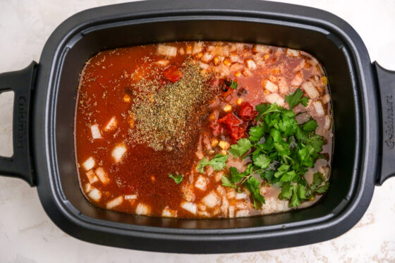 A slow cooker containing ingredients for slow cooker chicken enchilada soup: olive oil, yellow onion, garlic, chipotle peppers, adobo sauce, tomato sauce, roasted tomatoes, dried oregano, chili powder, cumin, salt, pepper, low sodium chicken broth, black beans, corn, and fresh cilantro.