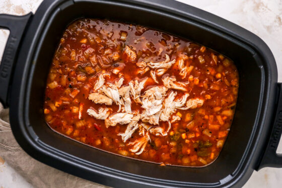 Shredded chicken added to a slow cooker containing ingredients for chicken enchilada soup.