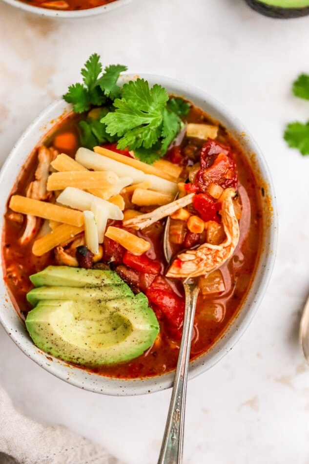 A spoon holding up a bite over a bowl of slow cooker enchilada soup. The soup has been topped with fresh avocado slices, cilantro and shredded cheese.