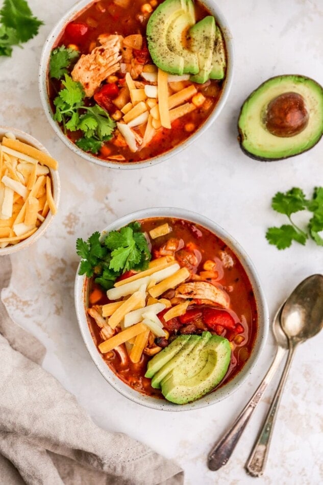 Two bowls of chicken enchilada soup, each topped with shredded cheese, fresh avocado slices and cilantro.