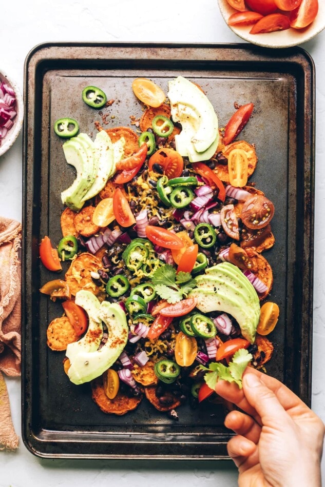 A sheet pan with sweet potato nachos and toppings. A hand is adding fresh chopped cilantro.