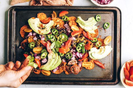 A sheet pan with sweet potato nachos and toppings. A hand is adding fresh chopped cilantro.