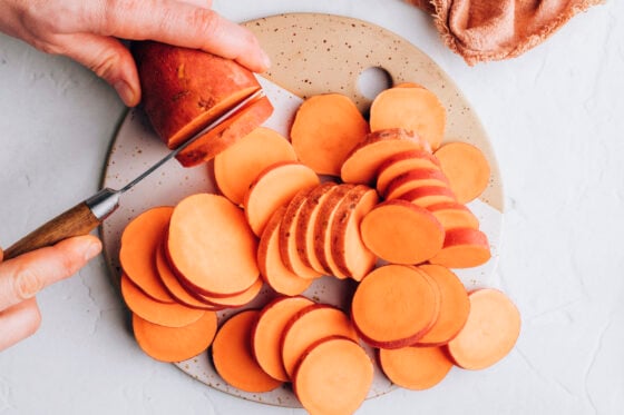 Two hands, one holding a sweet potato and the other using a knife to cut the sweet potato into thin, evenly sized rounds.