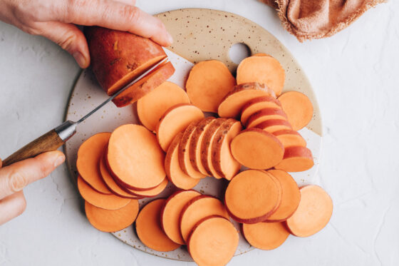 Two hands, one holding a sweet potato and the other using a knife to cut the sweet potato into thin, evenly sized rounds.