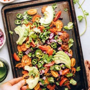 A hand scooping up a sweet potato round topped with black beans, cheese, avocado slices, and fresh cilantro.