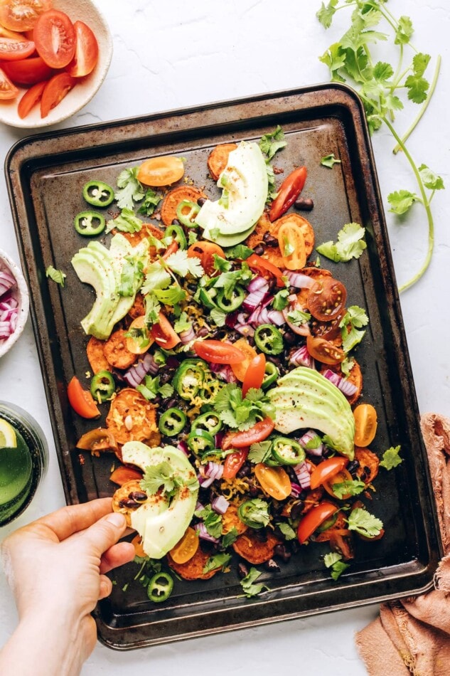 A hand scooping up a sweet potato round topped with black beans, cheese, avocado slices, and fresh cilantro.