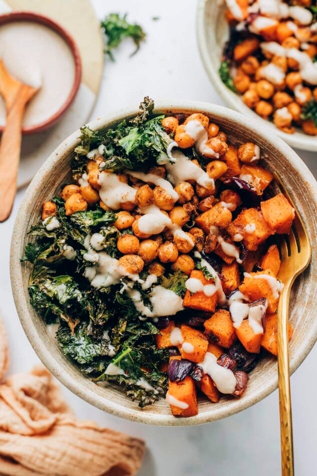 A bowl with roasted sweet potatoes and kale, drizzled with creamy white bean dressing. A gold fork rests in the bowl.