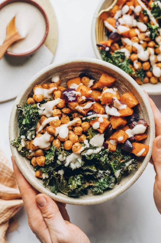 Hands holding up a bowl containing kale, chickpeas and roasted vegetables, drizzled with a creamy white bean dressing.