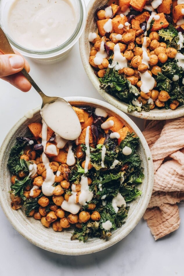 A gold spoon drizzling creamy white bean dressing on top of a bowl with roasted sweet potatoes and kale.