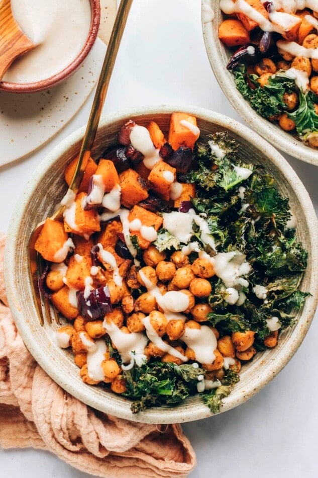 A vegan power bowl composed of kale, chickpeas and roasted veggies drizzled with a creamy white bean dressing. A gold fork rests in the bowl.