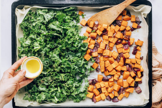 A parchment lined sheet pan. One half contains the chopped sweet potatoes and red onion, the other half contains the chopped kale. Olive oil is being poured overtop of the kale.
